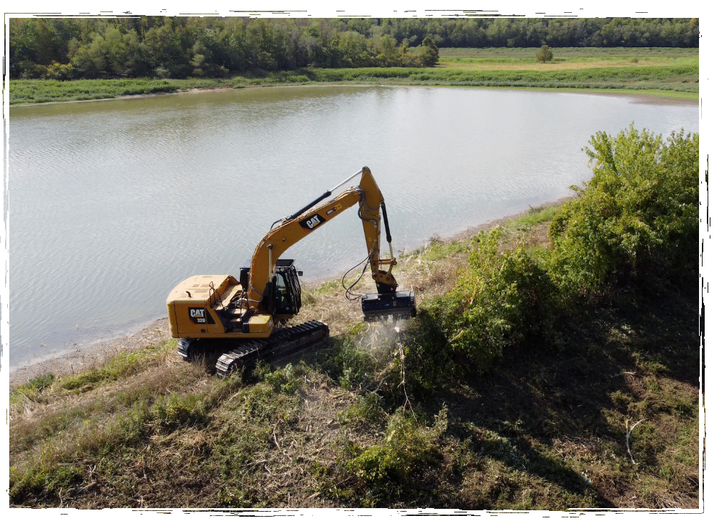Backhoe clearing brush and trees next to a lake